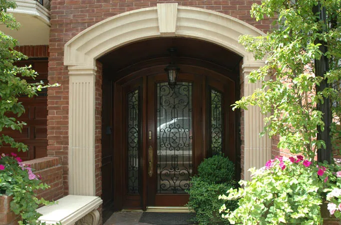 Arched wood entry door with iron-grille glass and sidelights, framed by flowering plants