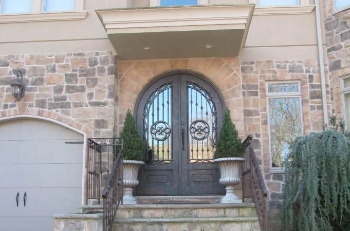 Arched iron double doors with scroll grilles between two potted evergreens